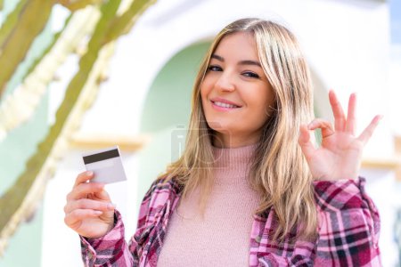 Foto de Joven mujer rumana bonita sosteniendo una tarjeta de crédito al aire libre mostrando signo ok con los dedos - Imagen libre de derechos