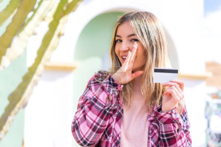 Téléchargez les photos : Jeune jolie roumaine tenant une carte de crédit en plein air chuchotant quelque chose - en image libre de droit