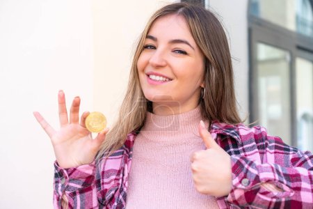 Photo for Young pretty Romanian woman holding a Bitcoin at outdoors with thumbs up because something good has happened - Royalty Free Image