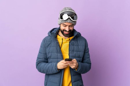 Téléchargez les photos : Skier homme avec des lunettes de snowboard sur fond violet isolé envoyer un message avec le mobile - en image libre de droit