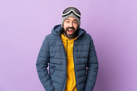 Téléchargez les photos : Skier homme avec des lunettes de snowboard sur fond violet isolé avec expression faciale surprise - en image libre de droit