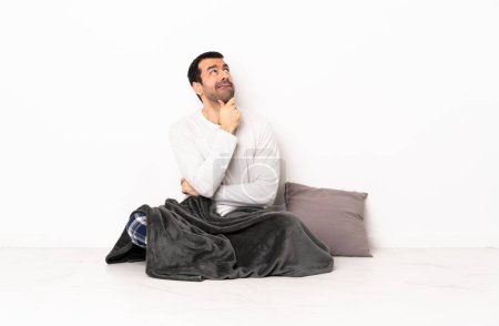 Téléchargez les photos : Homme caucasien en pyjama assis sur le sol à l'intérieur ayant des doutes et la pensée - en image libre de droit