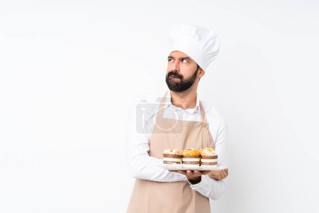 Foto de Joven sosteniendo pastel de magdalenas sobre fondo blanco aislado con expresión de cara confusa - Imagen libre de derechos