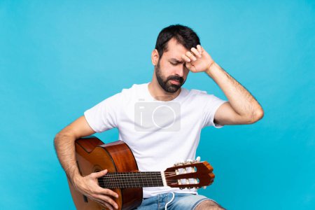 Foto de Joven con guitarra sobre fondo azul aislado con expresión cansada y enferma - Imagen libre de derechos