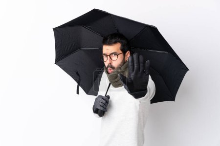 Foto de Caucásico hombre guapo con barba sosteniendo un paraguas sobre la pared blanca aislada haciendo gesto de parada y decepcionado - Imagen libre de derechos