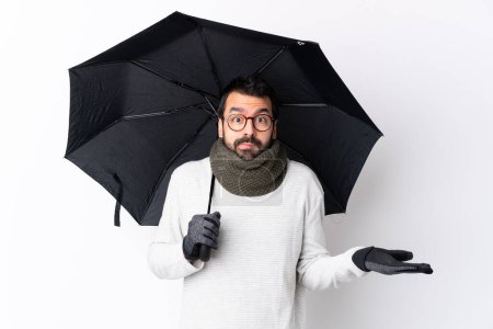 Foto de Caucásico guapo hombre con barba sosteniendo un paraguas sobre aislada pared blanca teniendo dudas mientras levanta las manos - Imagen libre de derechos