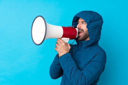 Photo for Man wearing winter jacket and holding a takeaway coffee over isolated blue background shouting through a megaphone - Royalty Free Image
