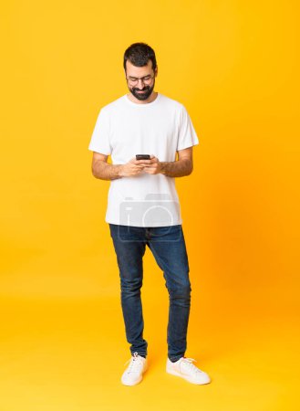 Photo for Full-length shot of man with beard over isolated yellow background sending a message with the mobile - Royalty Free Image