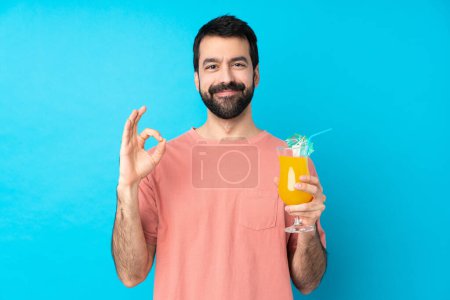 Téléchargez les photos : Jeune homme tenant un cocktail sur fond bleu isolé montrant un signe ok avec les doigts - en image libre de droit