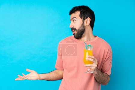 Téléchargez les photos : Jeune homme tenant un cocktail sur fond bleu isolé avec expression faciale surprise - en image libre de droit