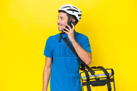Foto de Joven hombre guapo con mochila térmica sobre fondo amarillo aislado manteniendo una conversación con el teléfono móvil con alguien - Imagen libre de derechos