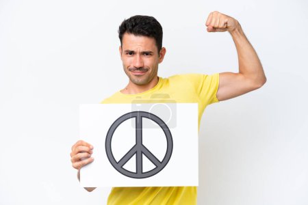 Téléchargez les photos : Jeune homme beau sur fond blanc isolé tenant une pancarte avec symbole de paix et faisant un geste fort - en image libre de droit