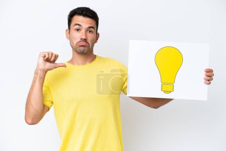 Photo for Young handsome man over isolated white background holding a placard with bulb icon with proud gesture - Royalty Free Image