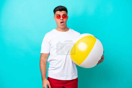 Téléchargez les photos : Jeune homme caucasien tenant une balle de plage isolée sur fond bleu levant les yeux et avec une expression surprise - en image libre de droit
