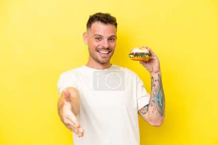 Téléchargez les photos : Jeune homme caucasien tenant un hamburger isolé sur fond jaune serrant la main pour conclure une bonne affaire - en image libre de droit
