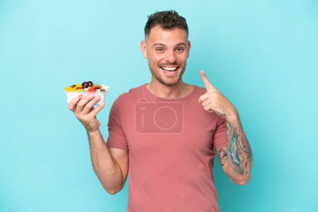 Téléchargez les photos : Jeune homme caucasien tenant un bol de fruits isolé sur fond bleu donnant un geste pouce vers le haut - en image libre de droit
