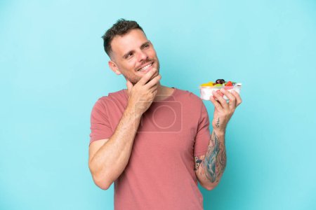 Foto de Joven hombre caucásico sosteniendo un tazón de fruta aislado sobre fondo azul mirando hacia arriba mientras sonríe - Imagen libre de derechos