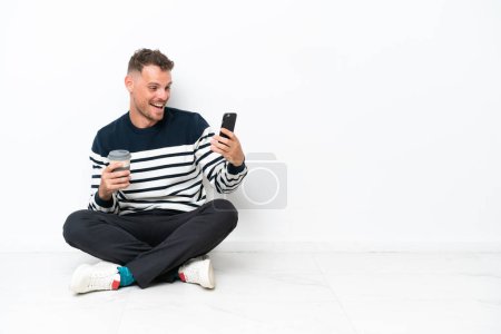 Téléchargez les photos : Jeune homme assis sur le sol isolé sur fond blanc tenant café à emporter et un mobile - en image libre de droit