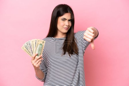 Téléchargez les photos : Young Brazilian woman taking a lot of money isolated on pink background showing thumb down with negative expression - en image libre de droit