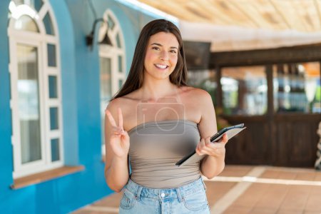 Téléchargez les photos : Jeune jolie Brésilienne tenant une tablette à l'extérieur souriant et montrant signe de victoire - en image libre de droit