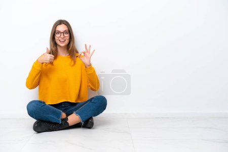 Téléchargez les photos : Jeune femme caucasienne assise sur le sol isolé sur fond blanc montrant signe ok et pouce geste vers le haut - en image libre de droit