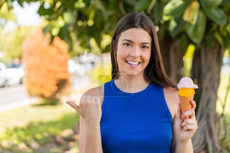 Téléchargez les photos : Jeune jolie Brésilienne avec une glace cornet à l'extérieur pointant vers le côté pour présenter un produit - en image libre de droit