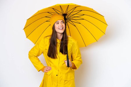 Téléchargez les photos : Jeune femme brésilienne avec manteau imperméable et parapluie isolé sur fond blanc posant avec les bras à la hanche et souriant - en image libre de droit