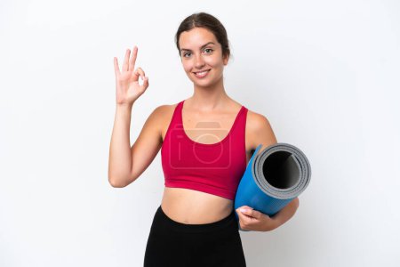 Foto de Young sport caucasian woman going to yoga classes while holding a mat isolated on white background showing ok sign with fingers - Imagen libre de derechos