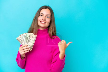 Téléchargez les photos : Jeune femme caucasienne prenant beaucoup d'argent isolé sur fond bleu pointant vers le côté pour présenter un produit - en image libre de droit