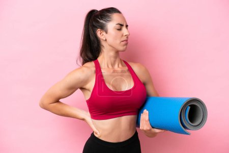 Téléchargez les photos : Jeune femme caucasienne sportive allant à des cours de yoga tout en tenant un tapis isolé sur fond rose souffrant de maux de dos pour avoir fait un effort - en image libre de droit