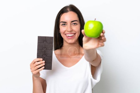 Jeune femme caucasienne isolée sur fond blanc prenant un comprimé de chocolat dans une main et une pomme dans l'autre