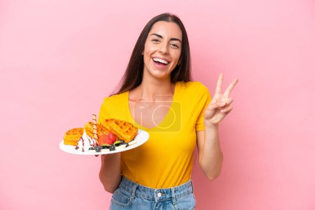 Photo for Young caucasian woman holding waffles isolated on pink background smiling and showing victory sign - Royalty Free Image