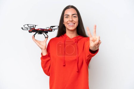 Téléchargez les photos : Jeune femme caucasienne tenant un drone isolé sur fond blanc souriant et montrant signe de victoire - en image libre de droit