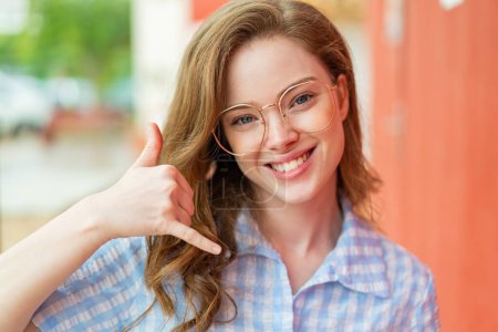 Téléchargez les photos : Jeune rousse femme à l'extérieur avec des lunettes et faire geste de téléphone - en image libre de droit