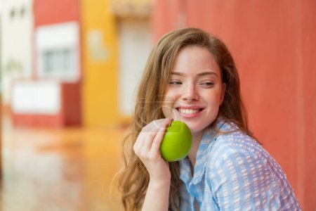 Téléchargez les photos : Jeune rousse femme à l'extérieur tenant une pomme avec une expression heureuse - en image libre de droit
