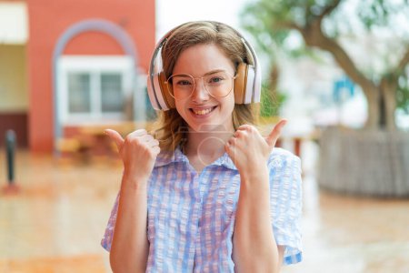Téléchargez les photos : Jeune rousse fille écouteurs à l'extérieur avec les pouces vers le haut geste et sourire - en image libre de droit