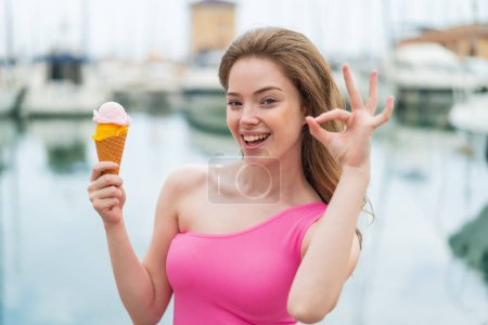 Téléchargez les photos : Jeune rousse femme avec une crème glacée cornet à l'extérieur montrant signe ok avec les doigts - en image libre de droit