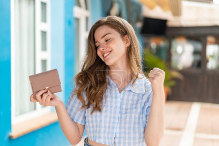 Téléchargez les photos : Jeune rousse femme tenant un portefeuille en plein air célébrant une victoire - en image libre de droit