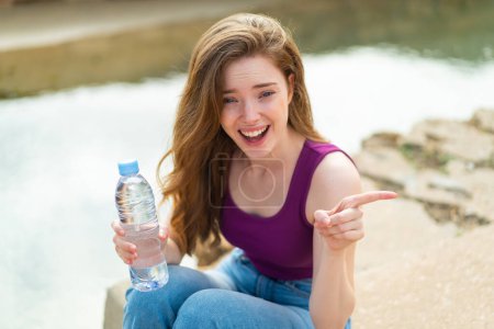 Téléchargez les photos : Jeune rousse femme avec une bouteille d'eau à l'extérieur surpris et pointant du doigt vers le côté - en image libre de droit