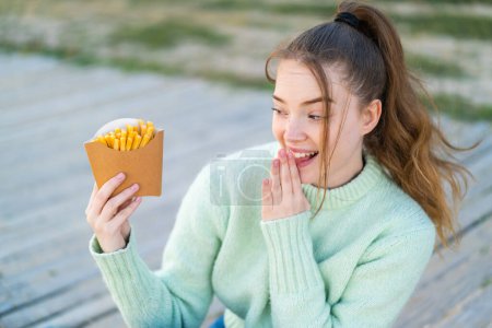 Téléchargez les photos : Jeune jolie fille tenant des frites à l'extérieur avec surprise et expression faciale choquée - en image libre de droit
