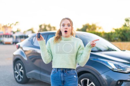 Téléchargez les photos : Jeune jolie fille tenant des clés de voiture à l'extérieur surpris et pointant côté - en image libre de droit