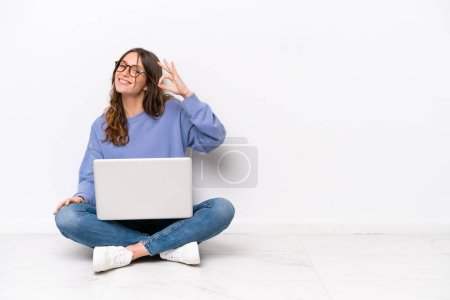 Foto de Young caucasian woman with a laptop sitting on the floor isolated on white background showing ok sign with fingers - Imagen libre de derechos