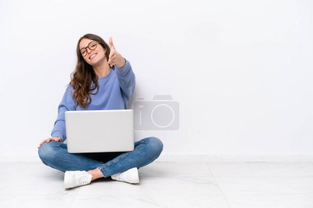 Téléchargez les photos : Jeune femme caucasienne avec un ordinateur portable assis sur le sol isolé sur fond blanc serrant la main pour fermer une bonne affaire - en image libre de droit