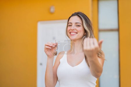 Téléchargez les photos : Jeune jolie femme tenant les clés de la maison à l'extérieur invitant à venir avec la main. Heureux que tu sois venu - en image libre de droit
