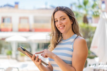 Téléchargez les photos : Jeune jolie femme à l'extérieur touchant l'écran de la tablette avec expression heureuse - en image libre de droit