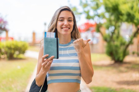Téléchargez les photos : Jeune jolie femme tenant un passeport à l'extérieur pointant vers le côté pour présenter un produit - en image libre de droit