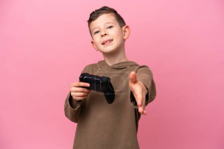 Téléchargez les photos : Petit garçon caucasien jouant avec un contrôleur de jeu vidéo isolé sur fond rose serrant la main pour conclure une bonne affaire - en image libre de droit