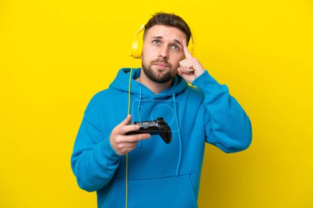 Téléchargez les photos : Jeune homme caucasien jouant avec un contrôleur de jeu vidéo isolé sur fond jaune ayant des doutes et la pensée - en image libre de droit