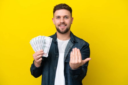 Photo for Young caucasian man taking a lot of money  isolated on yellow background inviting to come with hand. Happy that you came - Royalty Free Image