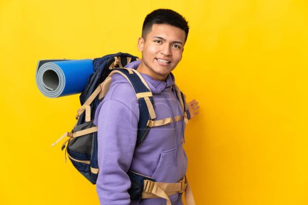 Foto de Joven montañero con una gran mochila aislada sobre fondo amarillo extendiendo las manos a un lado para invitar a venir - Imagen libre de derechos